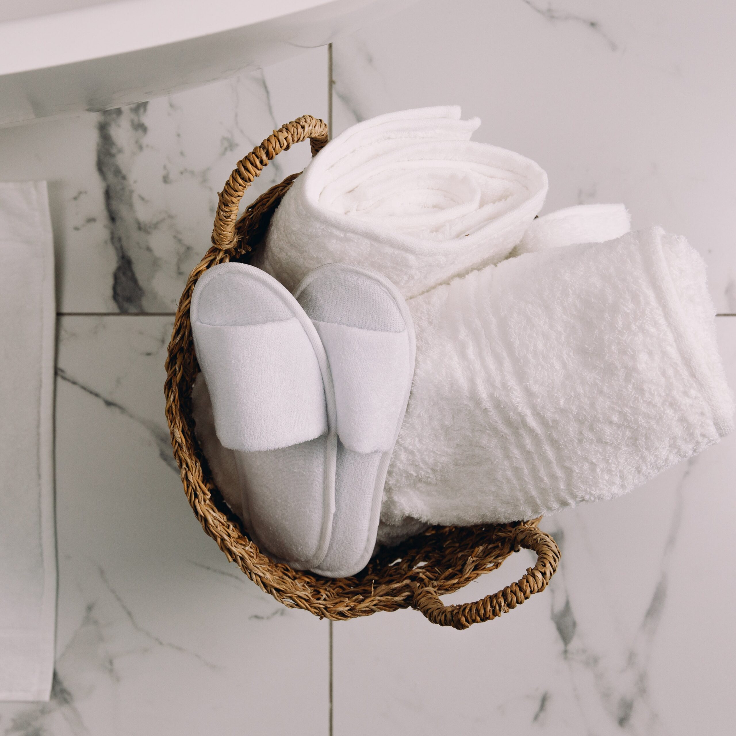 K1A0480-Edit White slippers and folded towels in a woven basket on a marble floor.