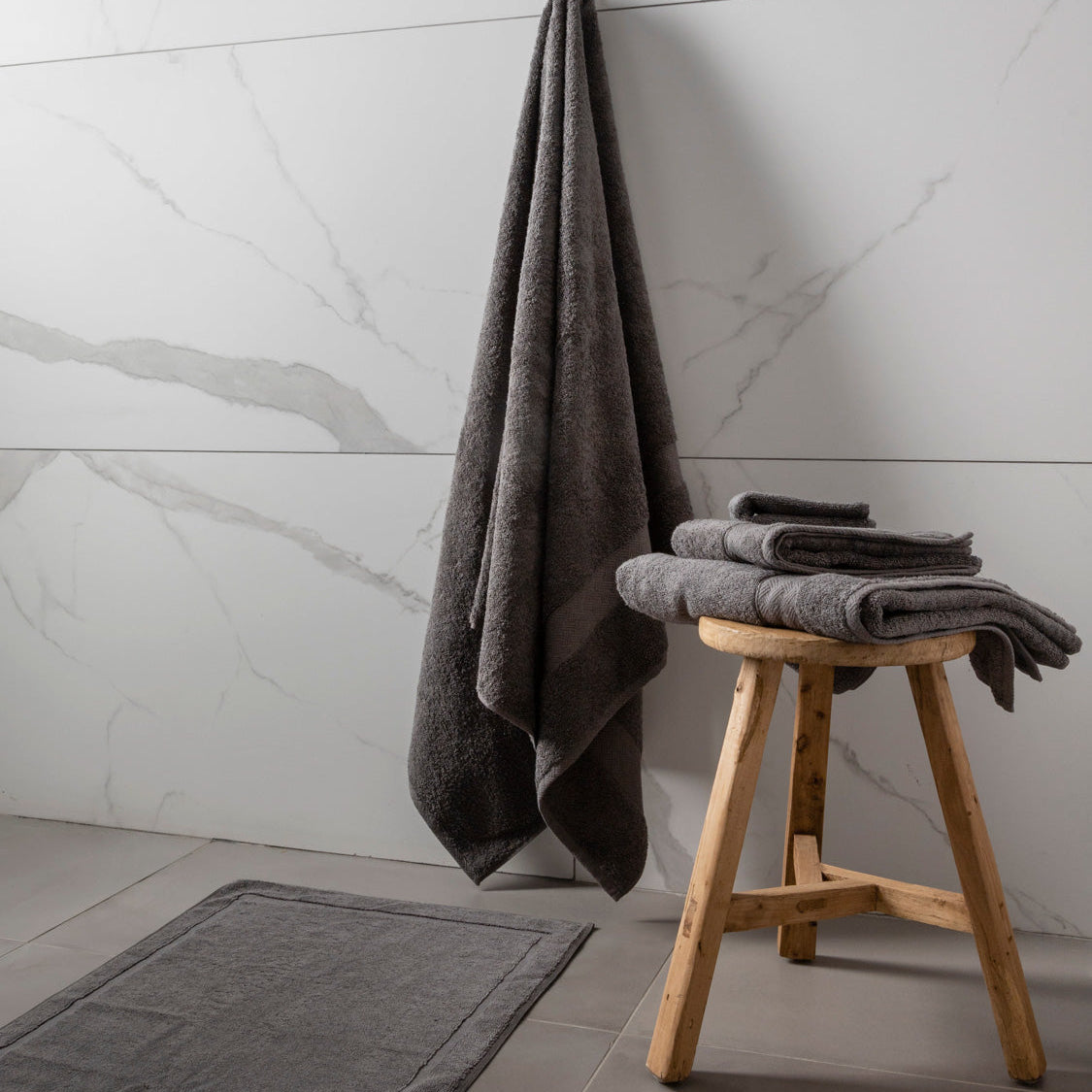 MG_9681_03b9d378-a2ef-455b-8299-5bc7019b6304-1 A set of bath towels in a bathroom setting, with one towel hanging on a wooden stool and the other two on a grey rug. The towels are in shades of grey and charcoal cloud, and the floor has a tiled design.